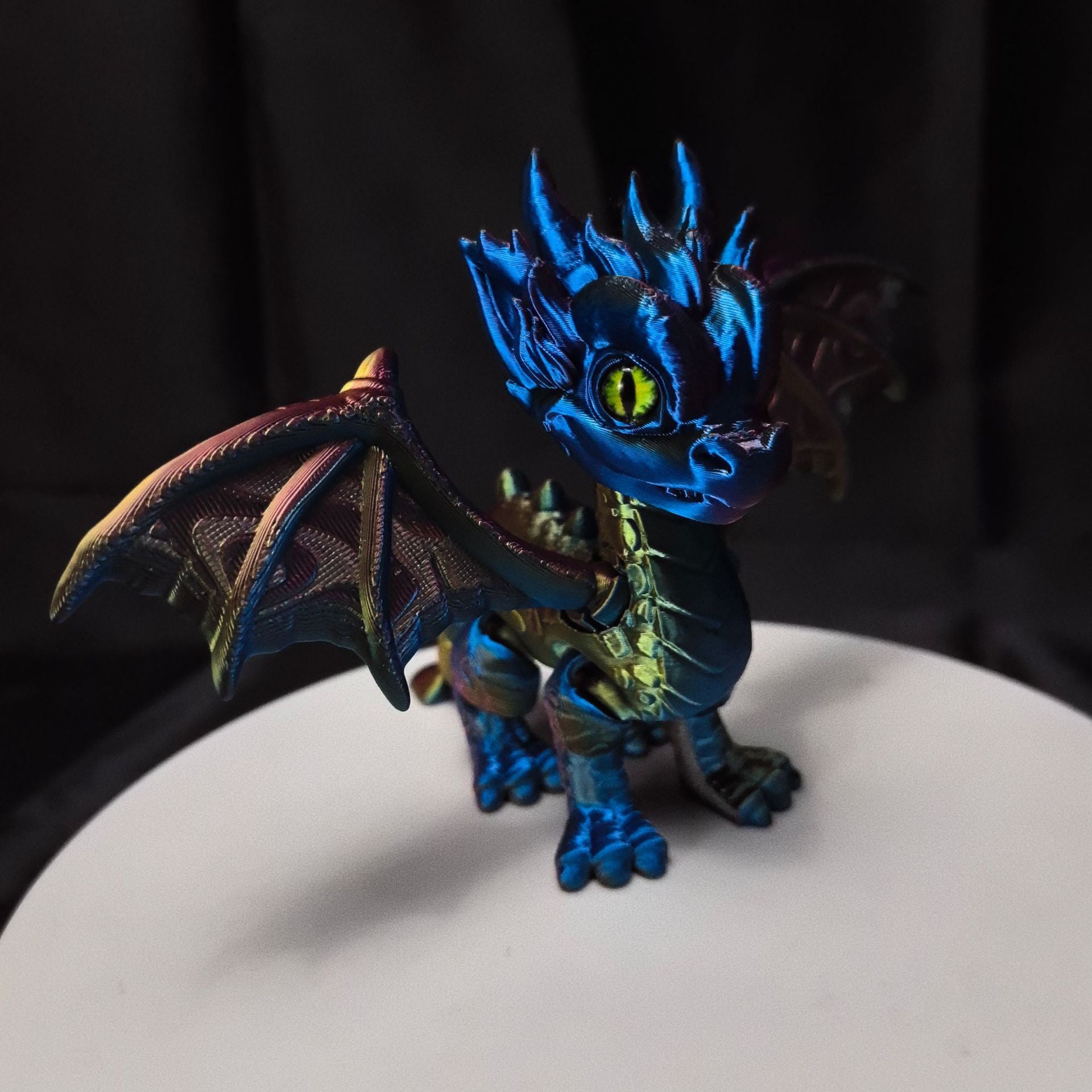 Blue green and purple iridescent posable baby dragon figurine standing on a white pedestal
