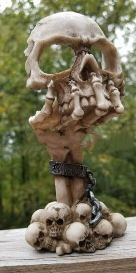 Skeletal hand gripping a carved skull aloft, chained at the wrist, rising from a pile of skulls